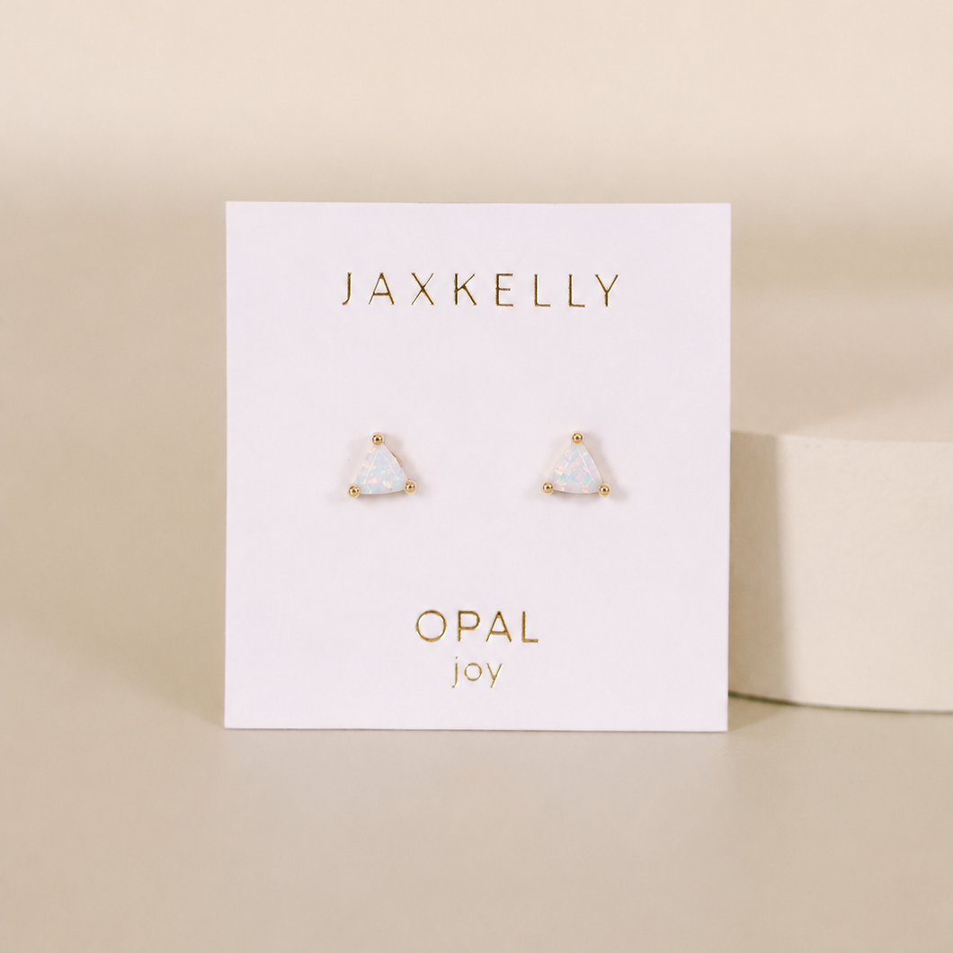 A pair of small, triangular lab-grown White Opal Mini Energy Gems - Joy studs by JaxKelly with 18k gold plating is shown on a white card labeled “JaxKelly” and “Opal joy” in gold, set against a neutral background.