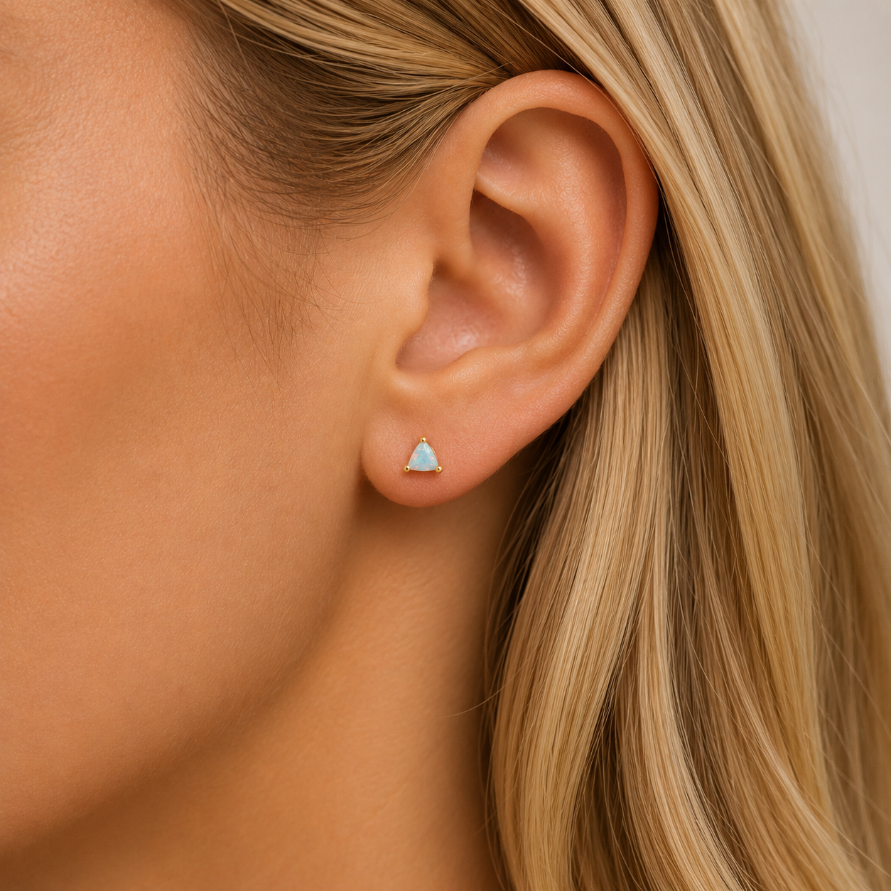 Close-up of a woman's ear with light skin and blonde hair, wearing JaxKelly's White Opal Mini Energy Gems - Joy stud earring set in 18k gold plating.