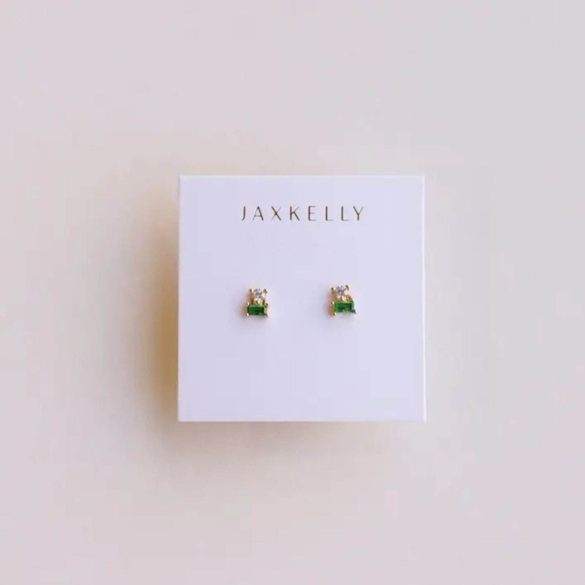 A pair of Double Stud Stack - Emerald CZ earrings by JaxKelly, gold plated with emerald baguette and clear round stones, displayed on a white JaxKelly card. These hypoallergenic studs provide stylish comfort for sensitive ears.