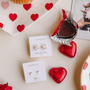 Two pairs of JaxKelly earrings—including the Tiny White Opal Heart—are displayed on a table with heart-shaped chocolates, a heart-patterned plate, and a partially unwrapped chocolate heart.