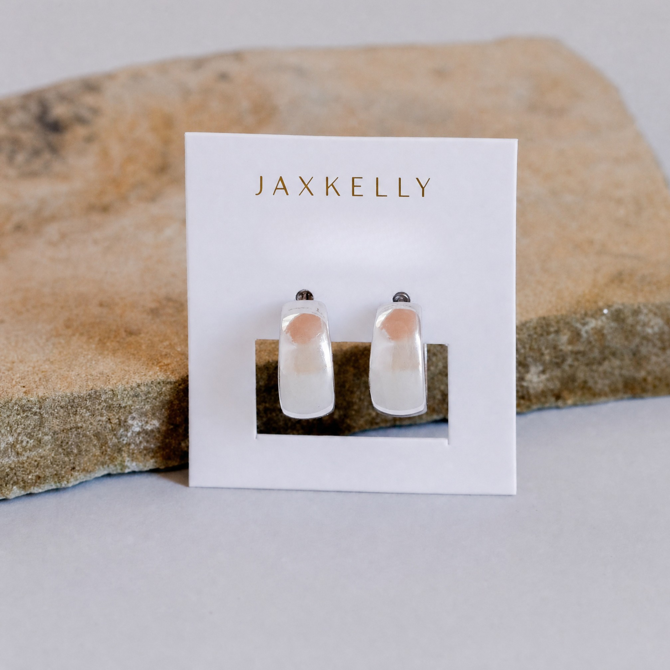 A pair of Silver Wide Hoop sterling silver earrings from JaxKelly, shown on a white card with gold lettering, propped against a textured stone background.