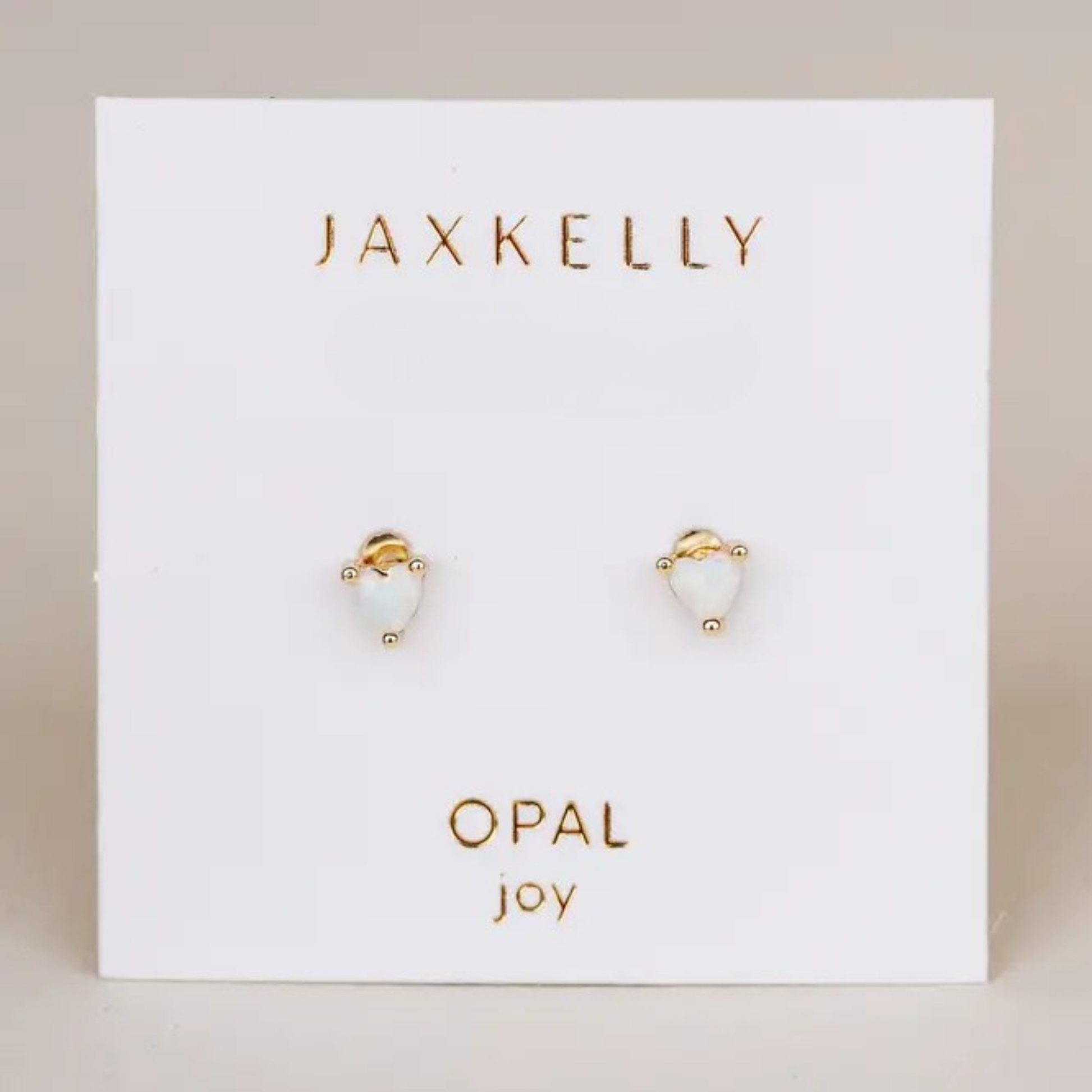 A pair of Tiny White Opal Heart studs by JaxKelly, featuring lab-grown opal in heart shapes, gold plated and displayed on a white card labeled “OPAL joy.”.