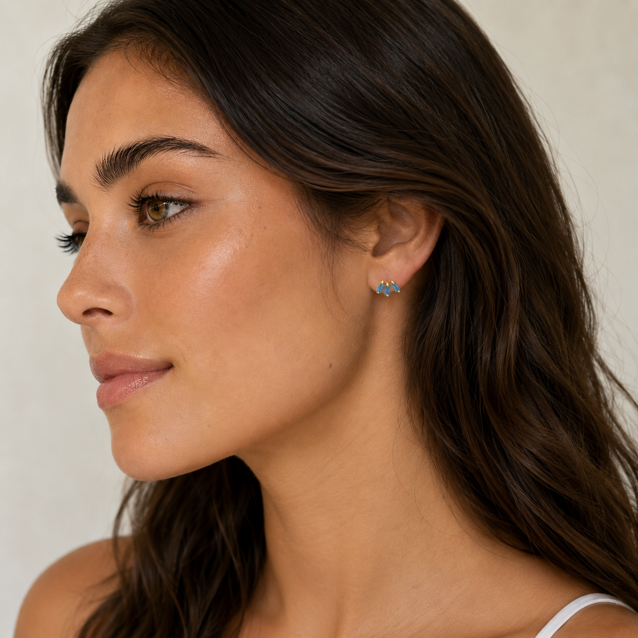 A woman with long brown hair and clear skin looks to the side, wearing JaxKelly’s Offset Trio - Fire Opal earrings and a white top, set against a neutral background.