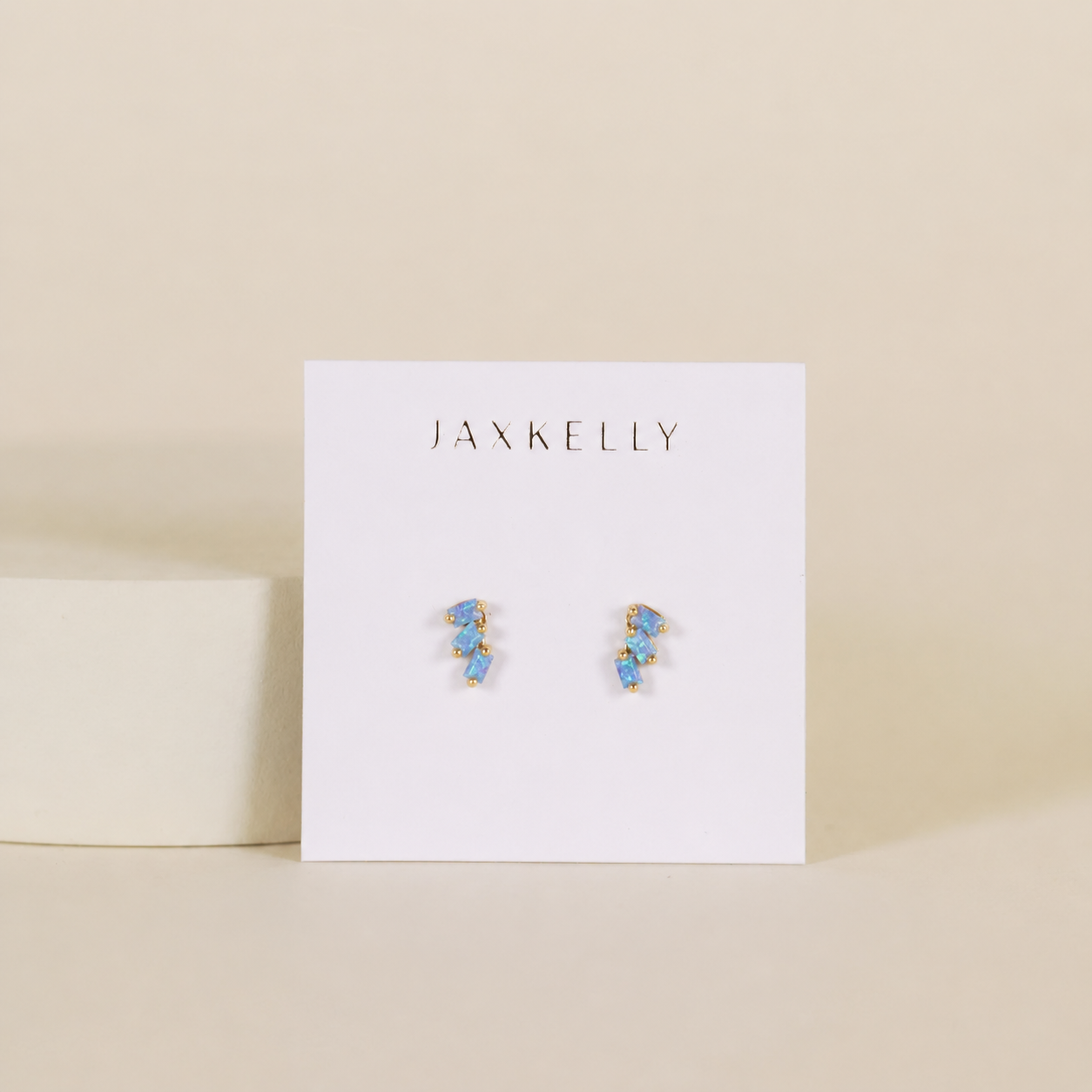 A pair of JaxKelly Offset Trio - Fire Opal gold-plated stud earrings with blue gemstones is displayed on a white JaxKelly card against a beige background.
