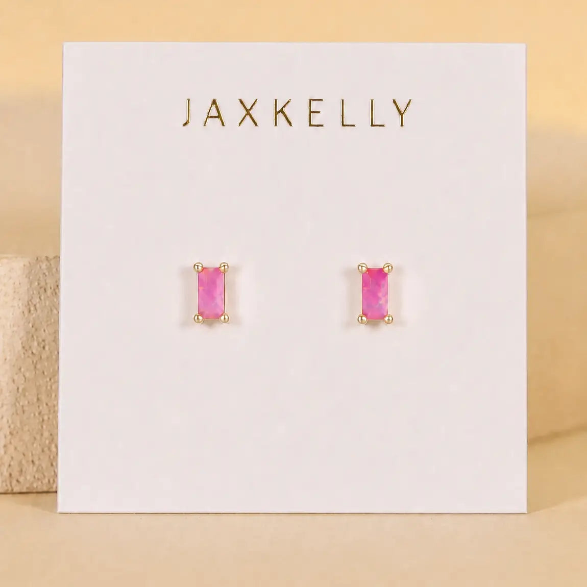 The JaxKelly Pink Opal Baguette earrings feature lab-grown pink opal gemstones in gold prongs, displayed on a white card with gold JaxKelly lettering. Hypoallergenic and perfect for sensitive ears.