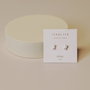 A pair of White Opal Baguette stud earrings are shown on a white card labeled JaxKelly Opal Joy, with a round beige object in the background on a neutral surface.