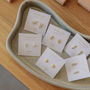 A tray displays seven pairs of JaxKelly White Opal Baguette stud earrings, 18k gold plated and lab-grown, on branded cards labeled "Opal joy," arranged neatly on a wooden surface.