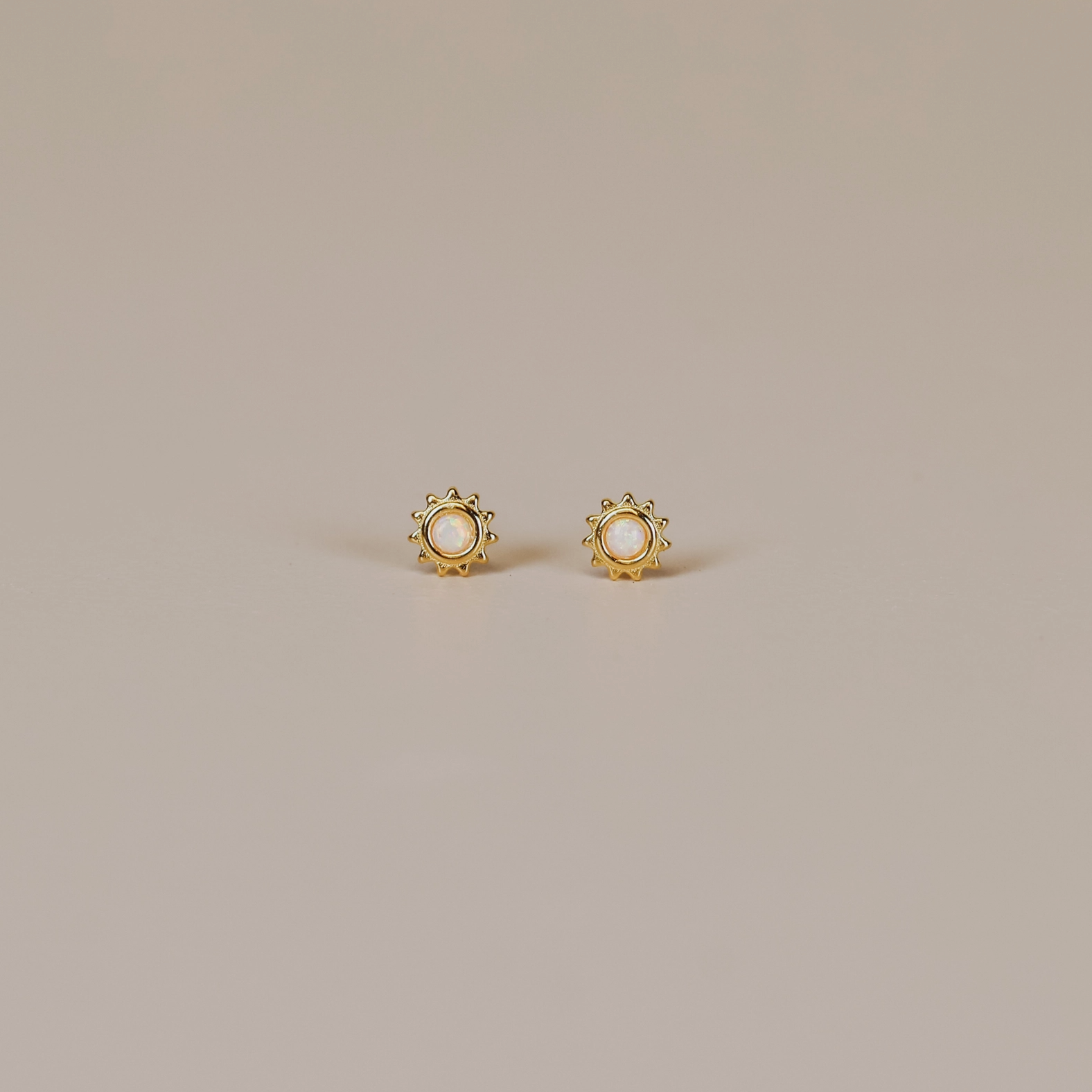 The White Opal Sun Stud features lab-grown white opal centers in a sun-inspired design, crafted with 18k gold plating and sterling silver posts, shown on a neutral light beige surface.