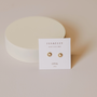 The White Opal Sun Stud earrings feature lab-grown white opals and 18k gold plating, shown on a Jax Kelly Opal Joy card with a round white object in the background on a light beige surface.