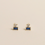 The Sapphire Double Stack earrings feature 18k gold plating with a small round CZ stone above a rectangular sapphire, shown against a plain light background.