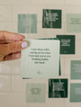 A hand with light purple nails holds an Abundance Shower Affirmations™ waterproof card reading, “I am okay with saying no to what does not serve me. Nothing holds me back,” under running water, with more cards on the tiled shower wall.