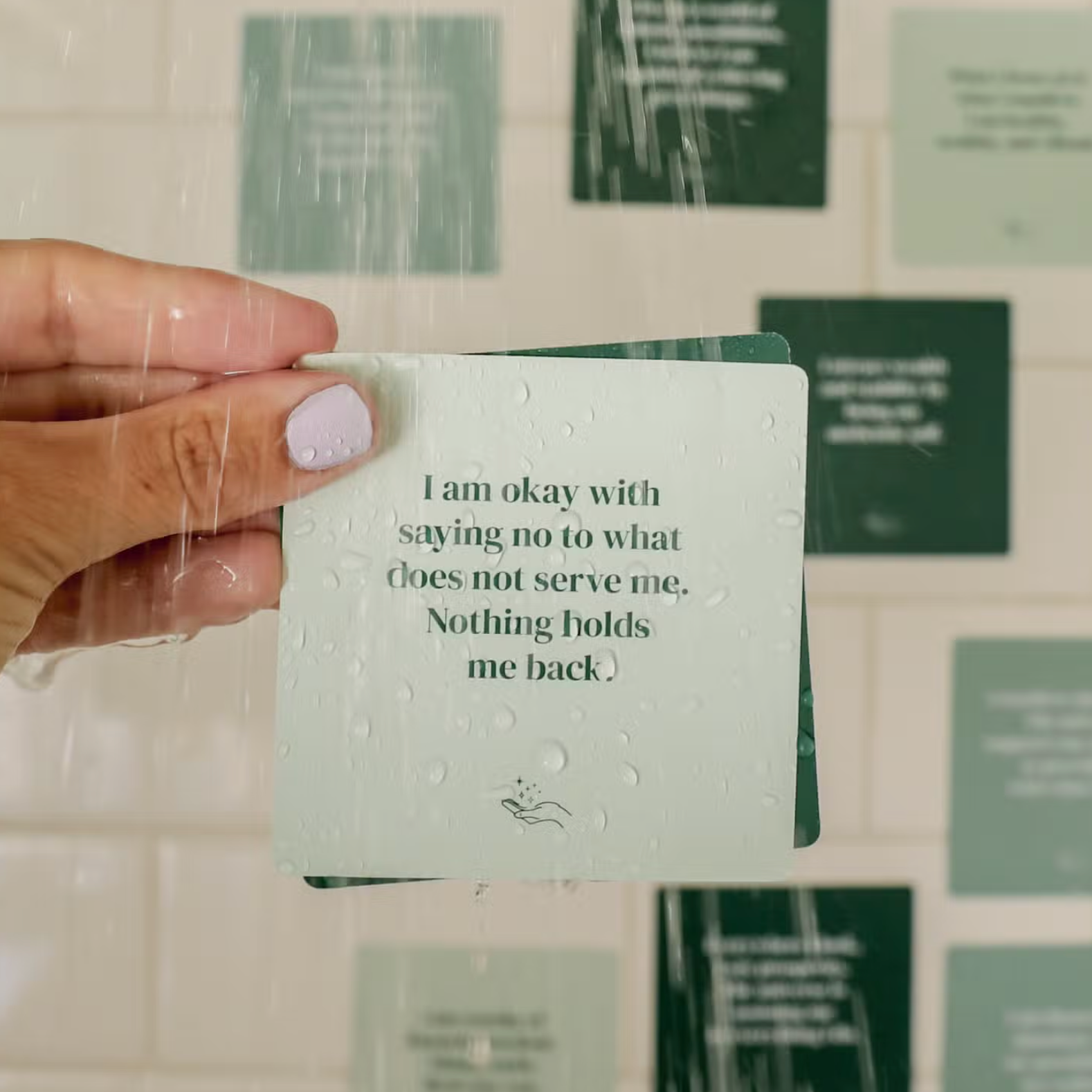 A hand with light purple nails holds an Abundance Shower Affirmations™ waterproof card reading, “I am okay with saying no to what does not serve me. Nothing holds me back,” under running water, with more cards on the tiled shower wall.