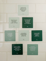 Nine Abundance Shower Affirmations™ cards with white text on green backgrounds are displayed in a grid on a white tiled wall. Each waterproof card features empowering quotes about self-worth and financial freedom.