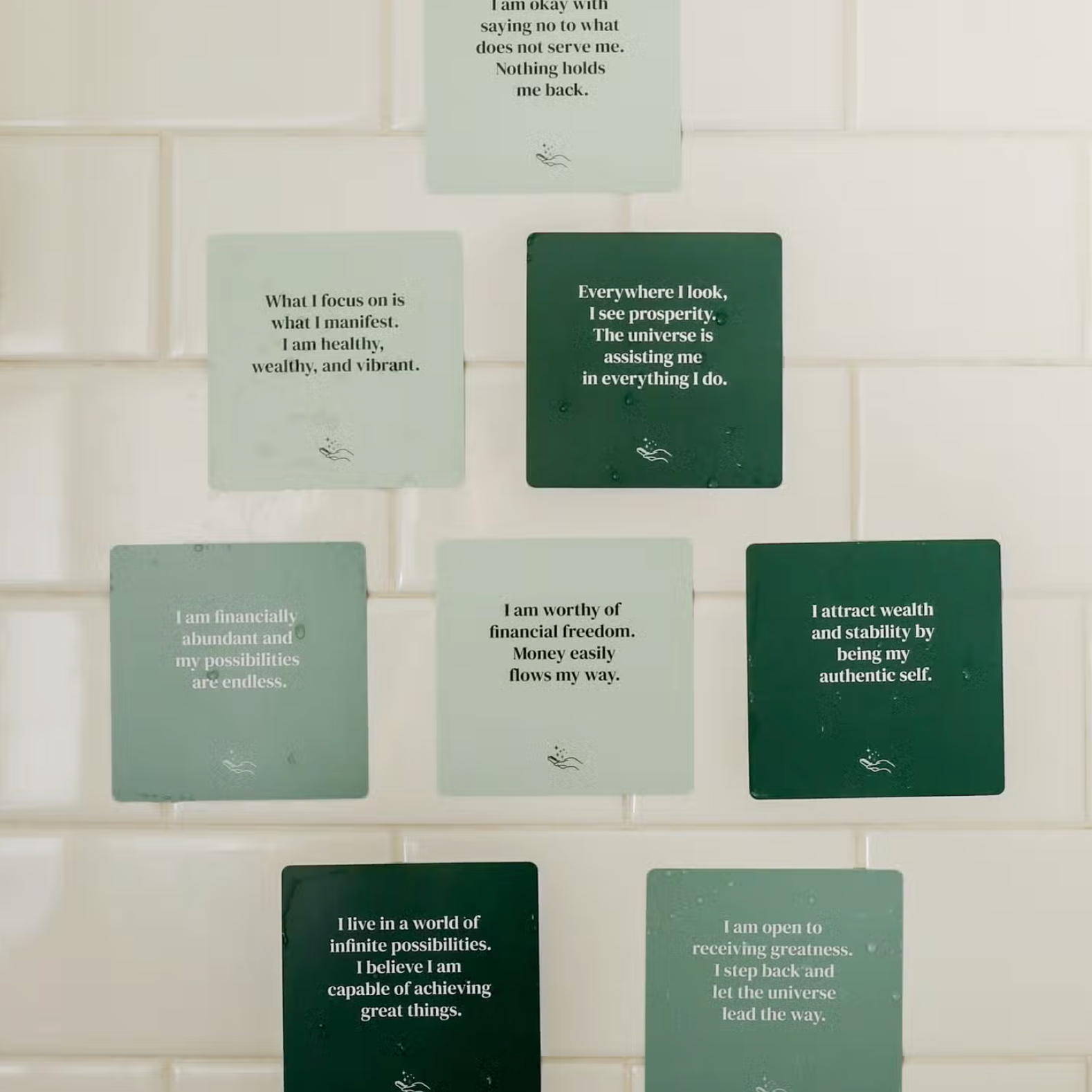 Nine Abundance Shower Affirmations™ cards with white text on green backgrounds are displayed in a grid on a white tiled wall. Each waterproof card features empowering quotes about self-worth and financial freedom.