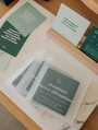 A set of green Abundance Shower Affirmations™ waterproof cards sits in a clear pouch on a wooden surface, with other motivational shower wall cards displayed nearby in a wooden holder.