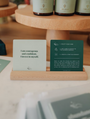 Two green waterproof Abundance Shower Affirmations™ cards on a wooden stand—one displays “I am courageous and confident. I invest in myself.”, the other gives instructions. Skincare products are blurred in the background.