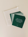 A stack of Abundance Shower Affirmations™ dark green waterproof cards, fanned out beside a translucent zippered pouch. The top card reads: "I have everything within me to succeed. Every day I am closer to my dreams.