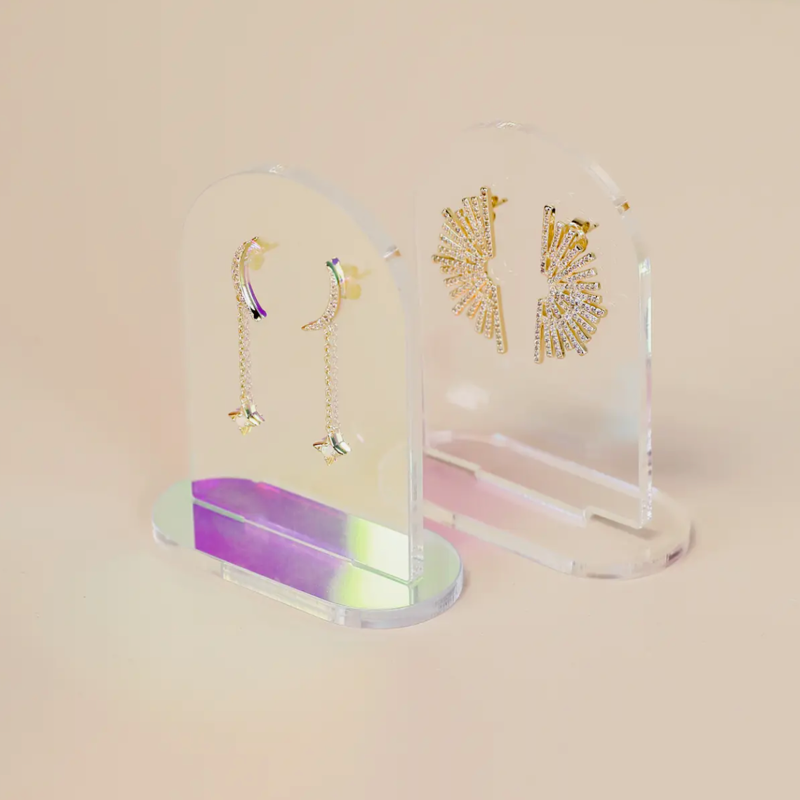 Two pairs of gold earrings are shown on Clear Earring Display Arch Solids: one with chain and small bow drops, the other with sparkly fan shapes. The display sits against a soft pastel beige background for a stylish retail look.
