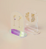 Two pairs of gold earrings are shown on Clear Earring Display Arch Solids: one with chain and small bow drops, the other with sparkly fan shapes. The display sits against a soft pastel beige background for a stylish retail look.