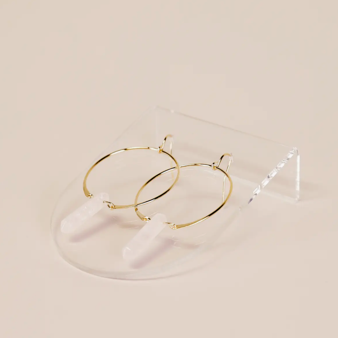 A pair of gold hoop earrings with clear quartz pendants is displayed on the Clear Earring Display Bent Large, a transparent curved acrylic stand, set against a light beige background.