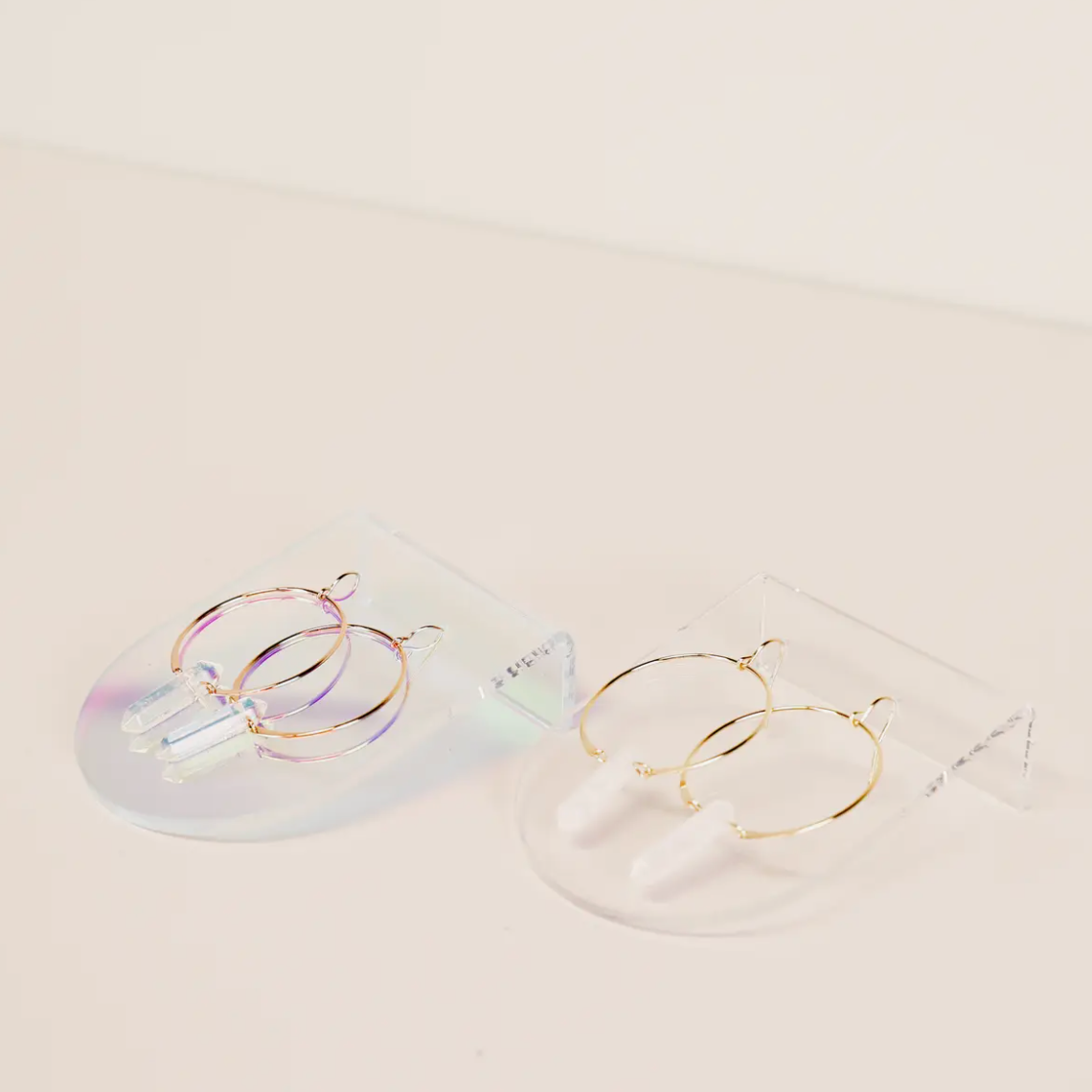 The Iridescent Earring Display Bent Large showcases two pairs of hoop earrings: iridescent hoops with crystal pendants on the left, and gold hoops with white cylindrical pendants on the right, all presented on minimalist acrylic stands.