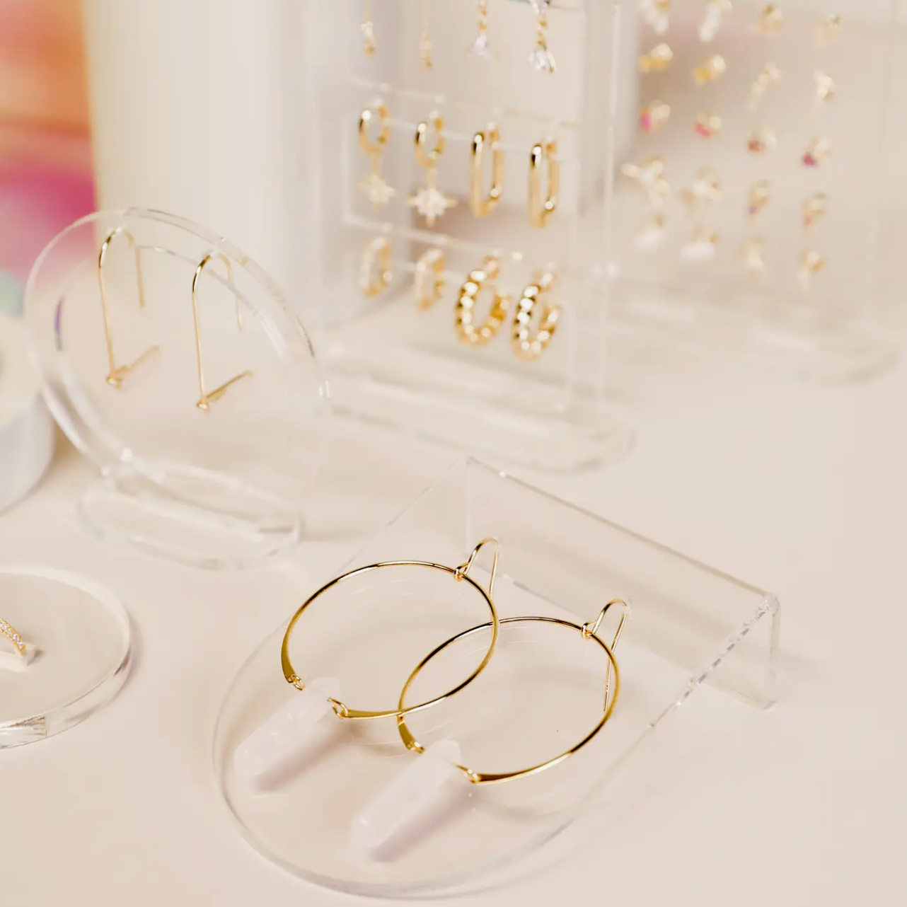 Gold hoop earrings with white stone pendants are featured on a minimalistic display, with other gold and jeweled earrings showcased on Clear Earring Display Bent Large acrylic holders in the background.