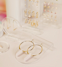 Gold hoop and dangling earrings are featured on the Iridescent Earring Display Bent Large, a minimalistic clear acrylic stand, with more delicate pairs in different shapes and styles softly blurred in the background.