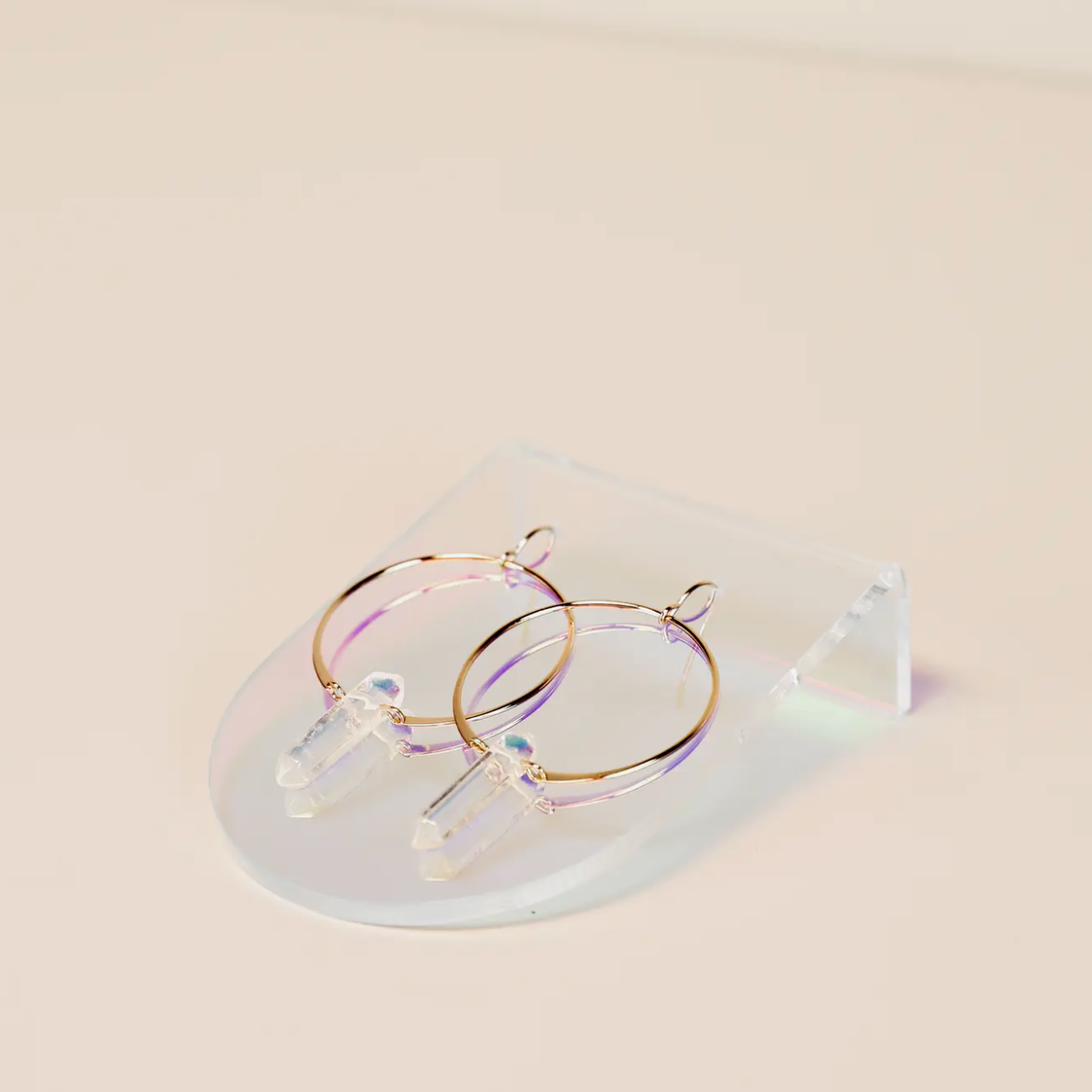 The Iridescent Earring Display Bent Large showcases a pair of gold hoop earrings with clear quartz pendants, set on a transparent iridescent stand against a light, neutral background.