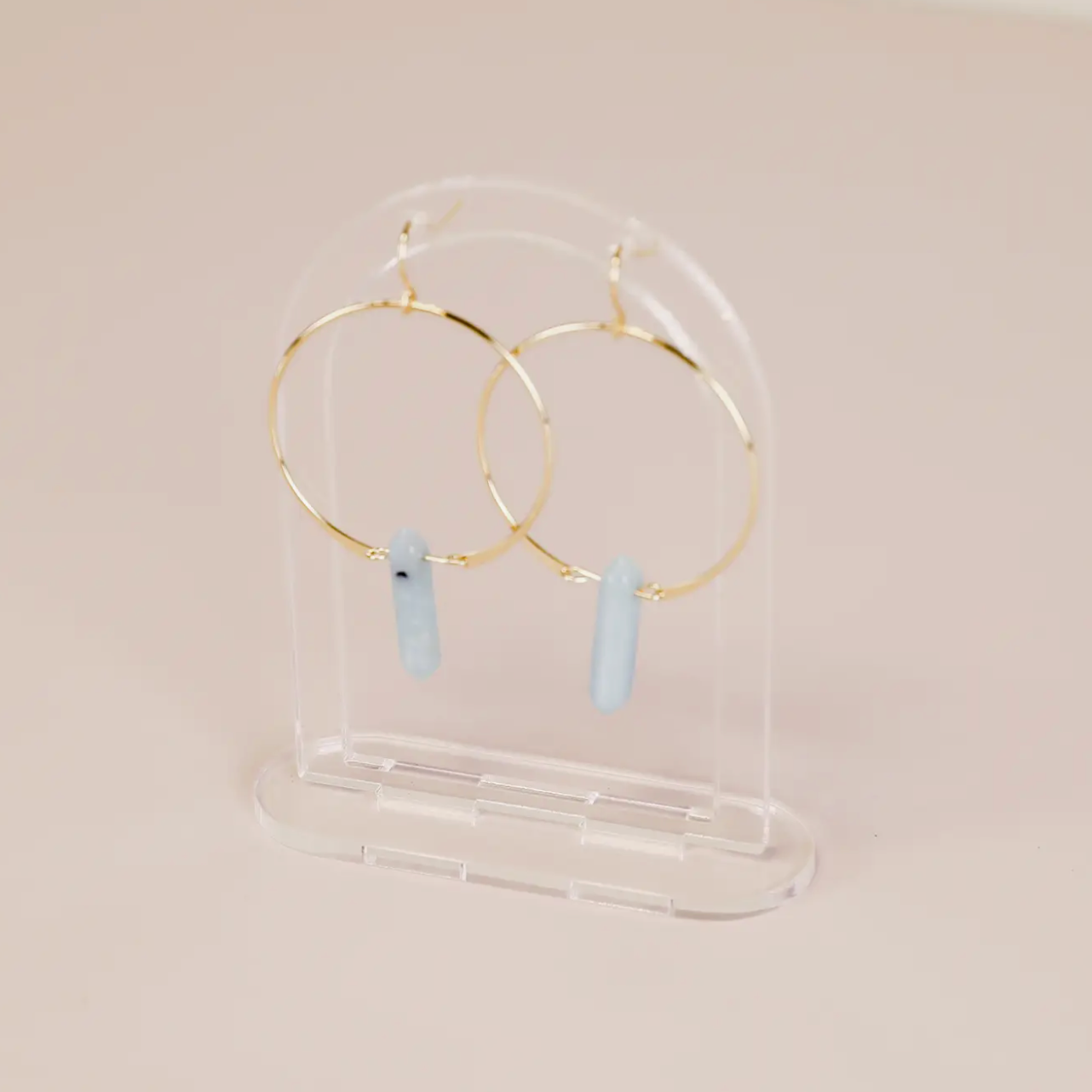 A pair of gold hoop earrings with light blue vertical stone pendants is displayed on the Clear Earring Display Hollow Arch, set against a pale pink background for a chic retail look.