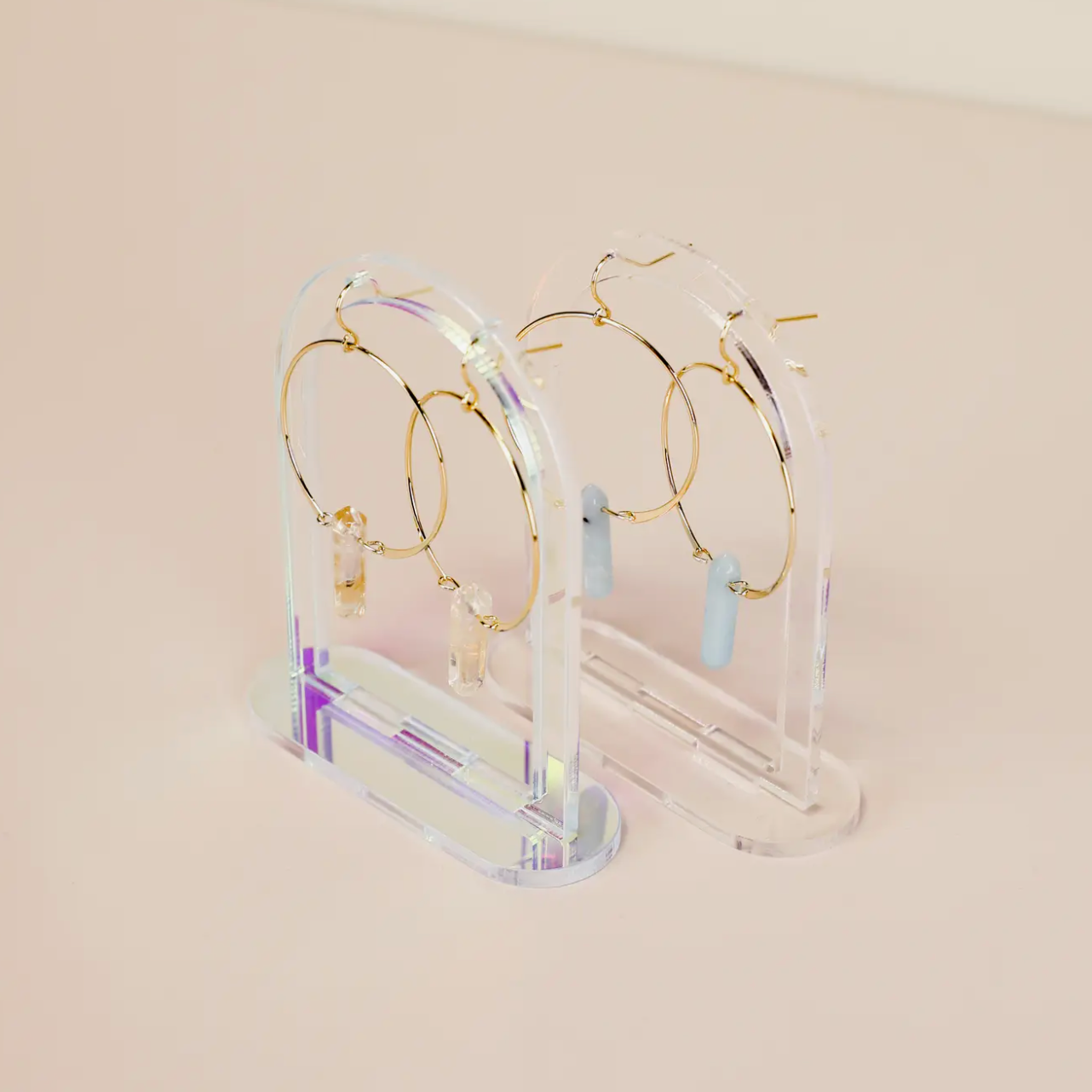 Two pairs of gold hoop earrings with gemstone dangles are showcased on Clear Earring Display Hollow Arch stands, highlighting a refined retail look against a soft beige background.