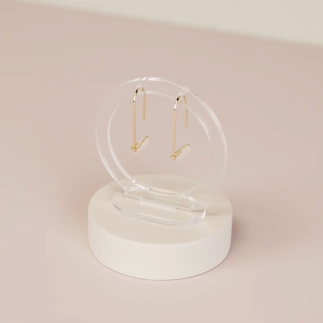 A pair of minimalistic gold earrings with slender vertical bars is elegantly displayed on a Clear Earring Display Hollow Circle with a white base, set against a soft beige background.