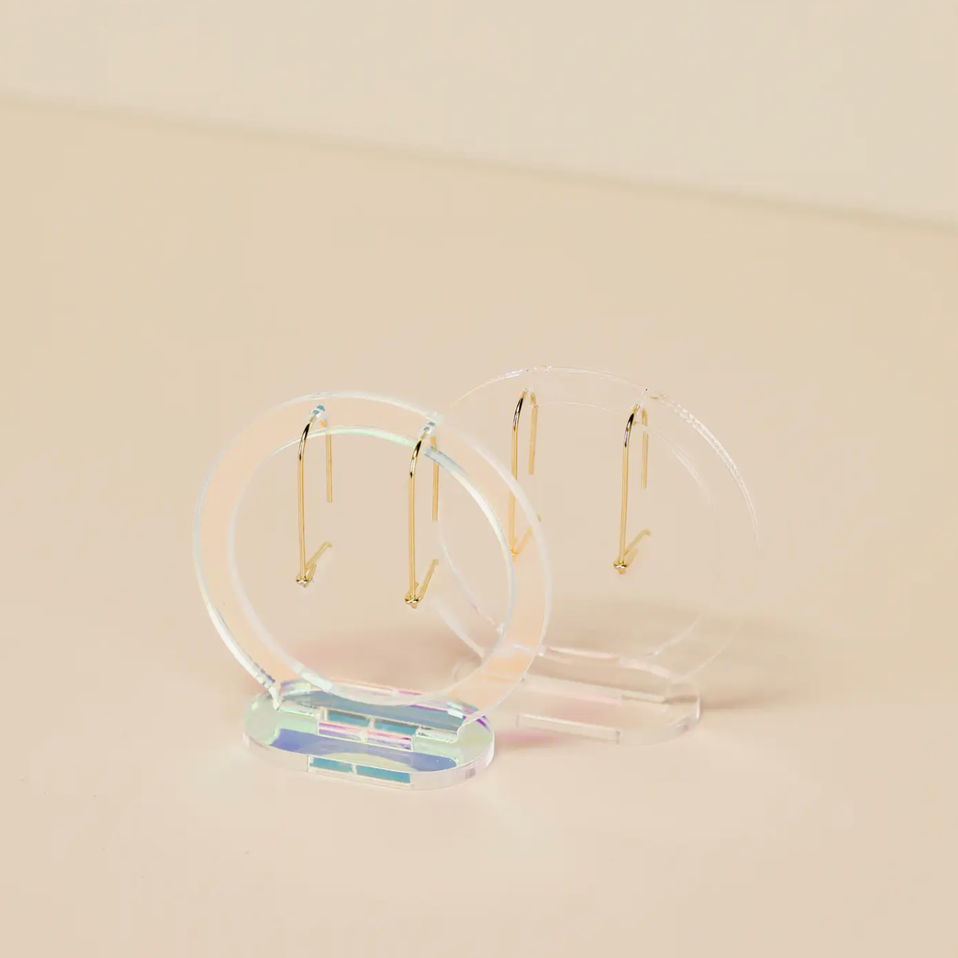 Two pairs of minimalistic gold hoop earrings displayed on Clear Earring Display Hollow Circle stands, made of clear, iridescent acrylic, set against a light beige background.