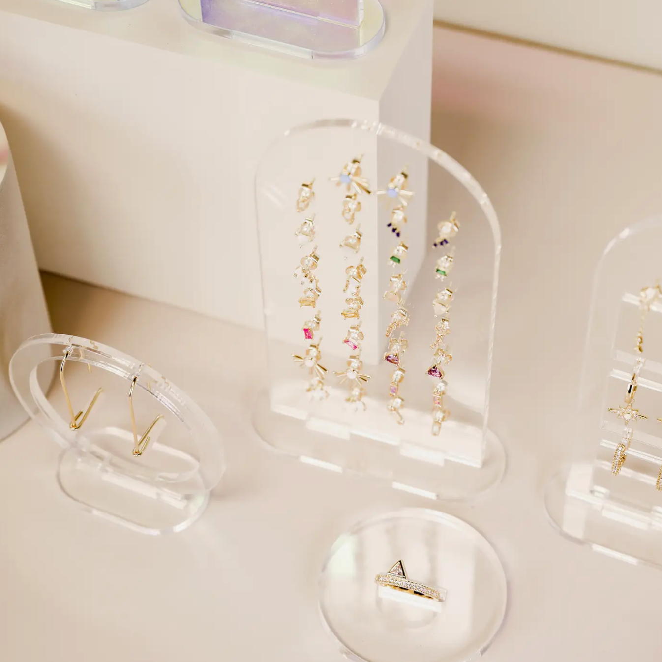 A retail display features gold jewelry, including colorful gemstone earrings on the Clear Earring Display Stud Arch, gold hoop earrings, and a triangular-design gold ring, all arranged on a light-colored surface inspired by JaxKelly.