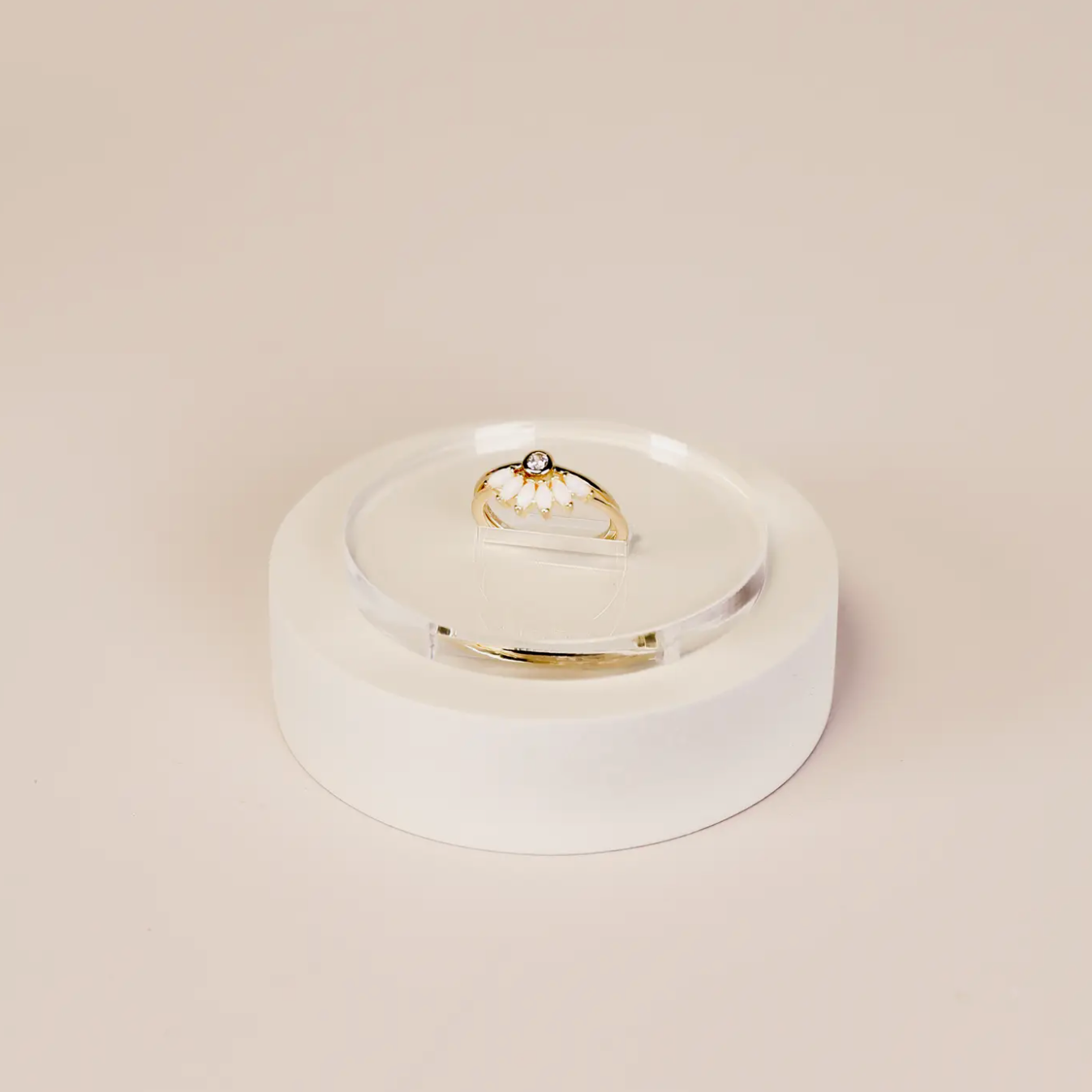 A gold ring with a white flower design and central gemstone is displayed on the Clear Ring Display Circle, set against a light beige background.