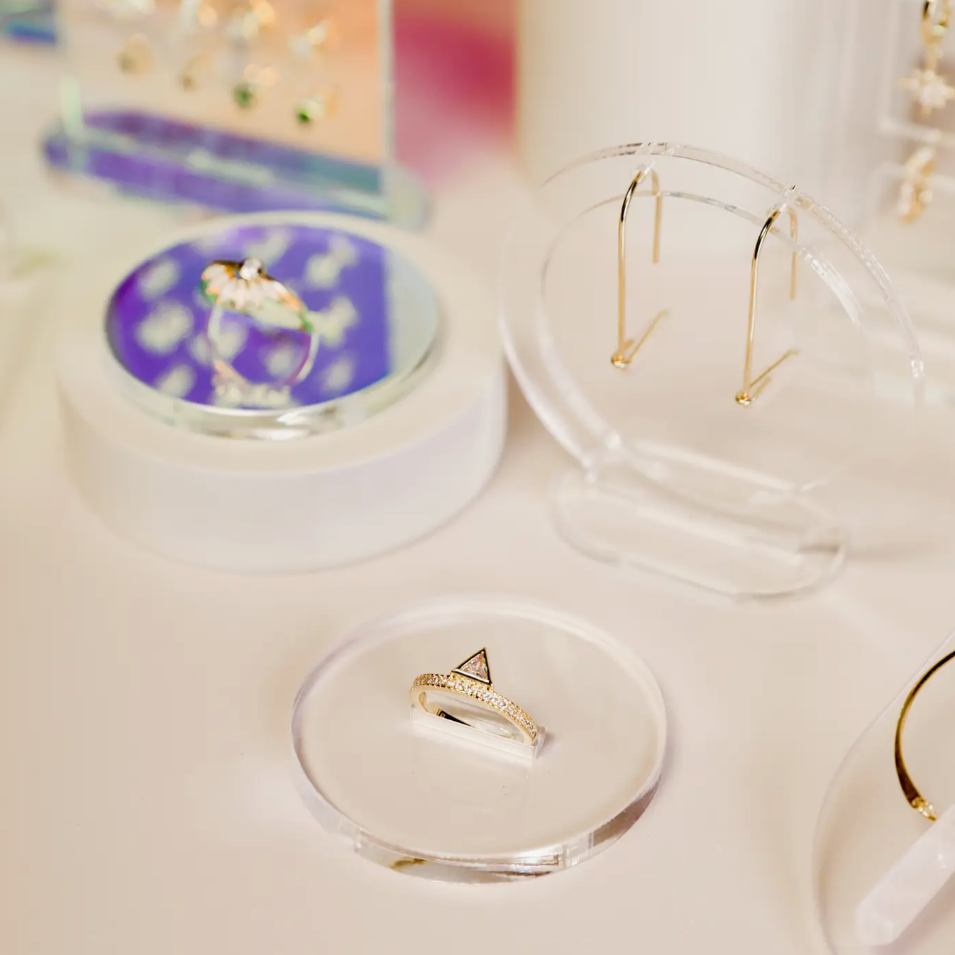 Elegant gold jewelry, including a statement ring on the Clear Ring Display Circle, thin hoop earrings, and other delicate pieces are arranged on a white surface with soft, pastel lighting.