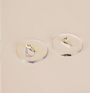 Two gold earrings rest separately on Clear Ring Display Circles against a pale beige background—one is a beaded hoop, the other a sleek curved stud—making them ideal for showcasing jewelry in a retail setting.