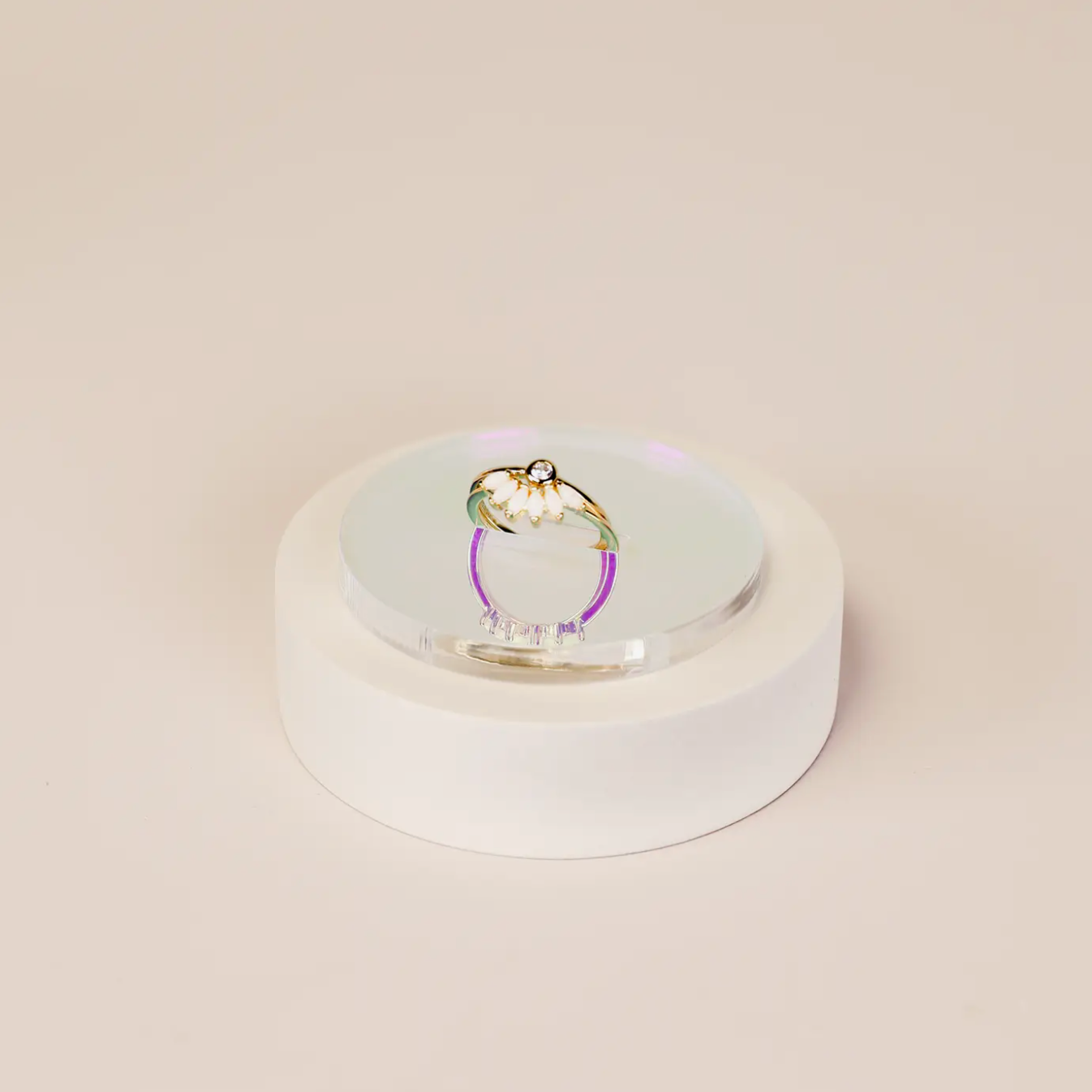 The Iridescent Ring Display Circle elegantly showcases a gold ring with a white flower design and central gemstone against a plain beige background, highlighting the refined style of JaxKelly displays.