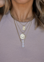 A woman wears a ribbed, light purple tank top and four JaxKelly gold necklaces—including crescent moon, rectangular pendant, sun and moon, and crystal—making the Mystery Necklace perfect for those who love a surprise accessory.