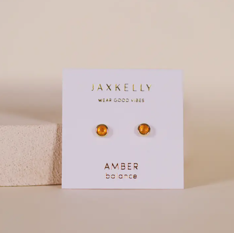 The Simple Stud Earring - Amber Gemstone features round amber studs with sterling silver posts, displayed on a white card that says "JAXKELLY Wear Good Vibes" and "AMBER balance," set against a neutral background.