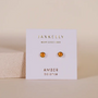 The Simple Stud Earring - Amber Gemstone features round amber studs with sterling silver posts, displayed on a white card that says "JAXKELLY Wear Good Vibes" and "AMBER balance," set against a neutral background.