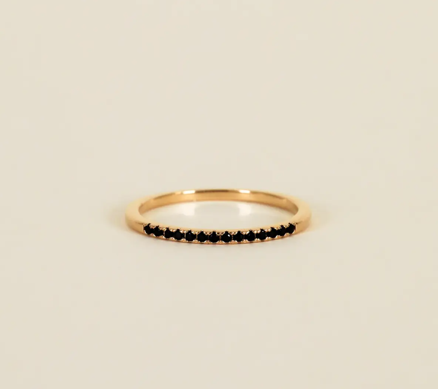The Half Pave Ring - Black features a slim gold band adorned with a row of small black cubic zirconia stones along the top, showcased on a plain beige background.