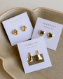 The Petite Gold Knot Earrings are displayed on a white JaxKelly card atop a beige ceramic dish, making them perfect for everyday wear.