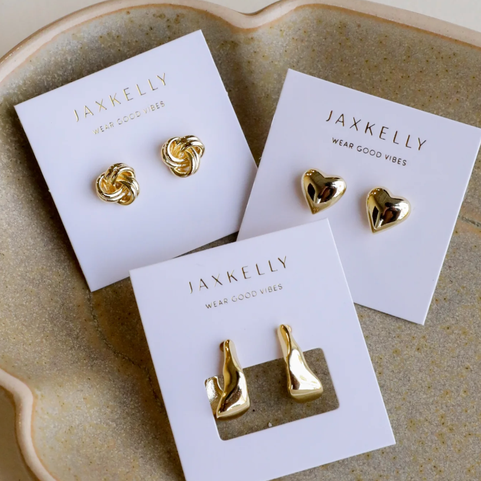 The Petite Gold Knot Earrings are displayed on a white JaxKelly card atop a beige ceramic dish, making them perfect for everyday wear.
