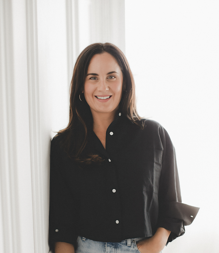 A woman with long dark hair, wearing a black button-up shirt and light jeans, stands indoors near a white wall, smiling at the camera with one hand in her pocket.