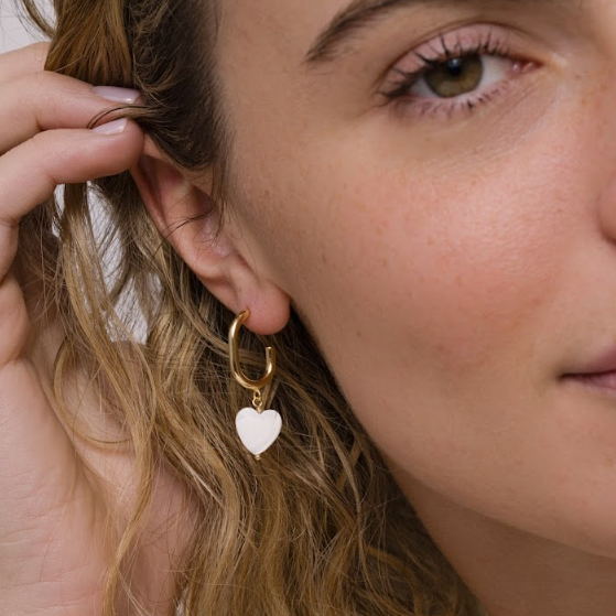 A close-up of a person with wavy blonde hair wearing JaxKelly Heart Drop - Mother of Pearl tarnish free gold hoop earrings featuring a white heart-shaped charm, gently touching their ear.