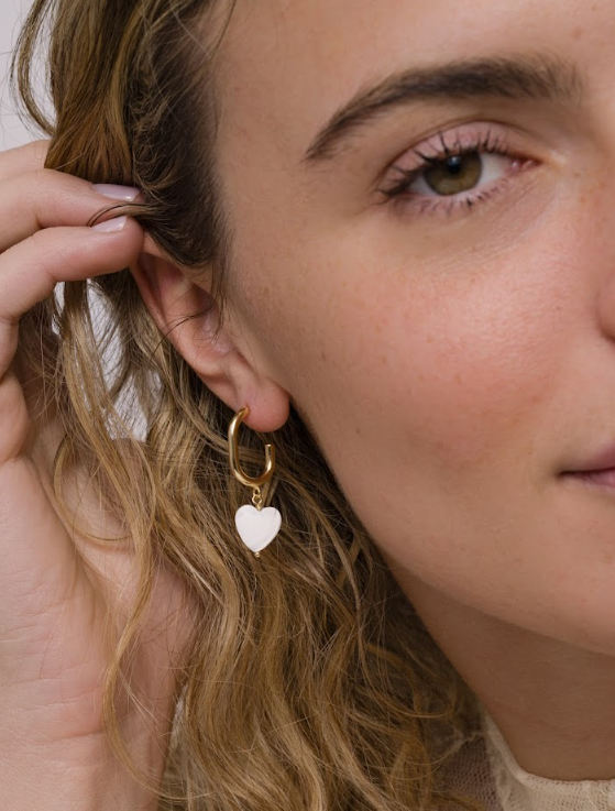 A close-up of a person with wavy blonde hair wearing JaxKelly Heart Drop - Mother of Pearl tarnish free gold hoop earrings featuring a white heart-shaped charm, gently touching their ear.
