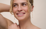 A young woman with light skin and blonde hair smiles, gazing to the side. She wears JaxKelly's Gemstone Chain Drop - Rose Quartz earrings with 18k gold plating, a gold ring, one arm raised and her other hand gently touching her shoulder.