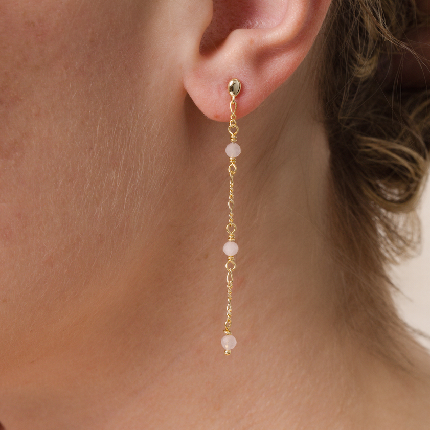 Close-up of a person wearing JaxKelly’s Gemstone Chain Drop - Rose Quartz earring, featuring three rose quartz beads on a delicate 18k gold-plated chain. The background is neutral, with the person's hair partially visible.