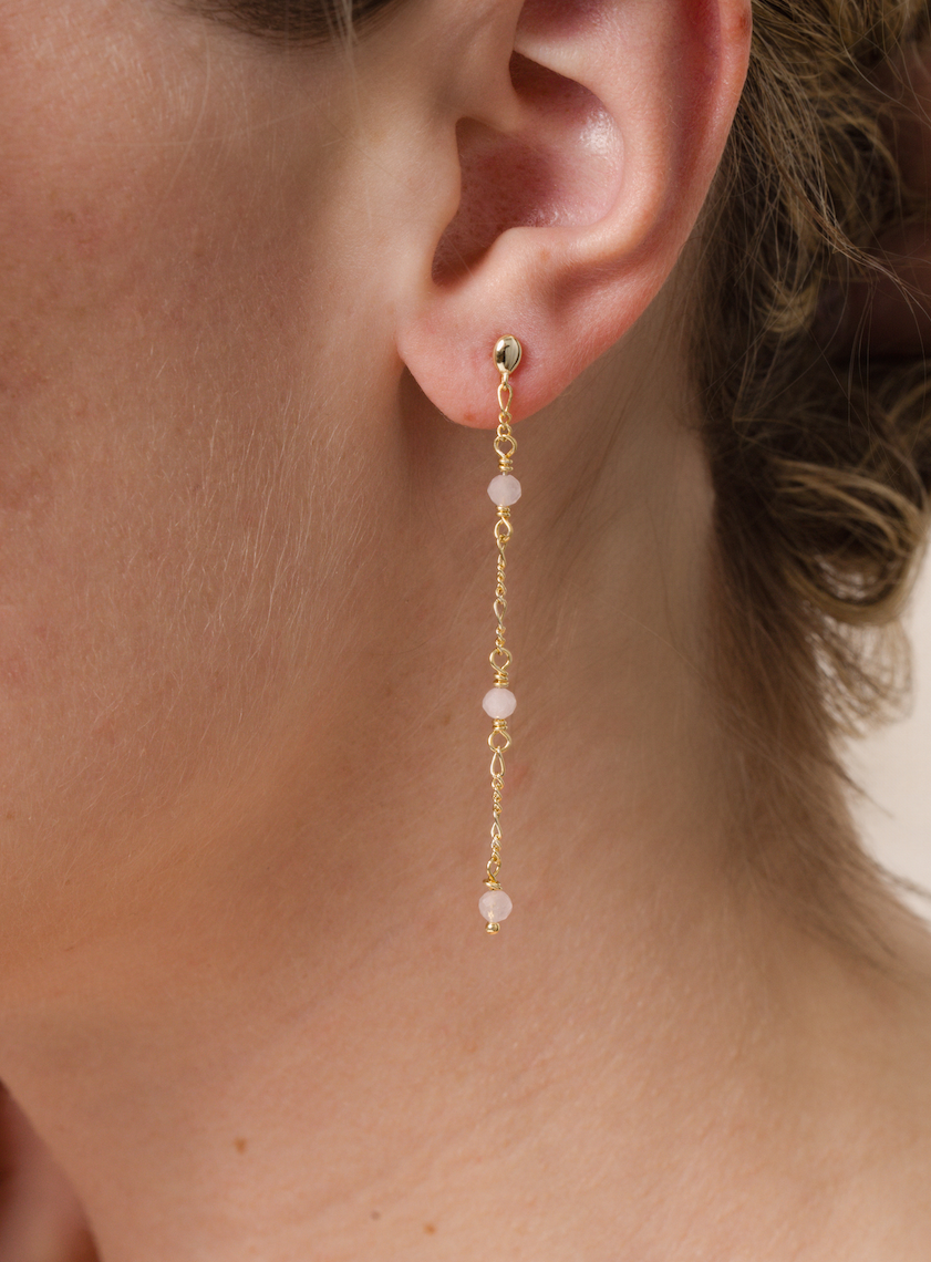 Close-up of a person wearing JaxKelly’s Gemstone Chain Drop - Rose Quartz earring, featuring three rose quartz beads on a delicate 18k gold-plated chain. The background is neutral, with the person's hair partially visible.