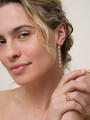 A woman with wavy blonde hair smiles softly, wearing JaxKelly's Multi Pearl Drop earrings and several 18k gold plated rings, posing with her hand near her face against a neutral background.